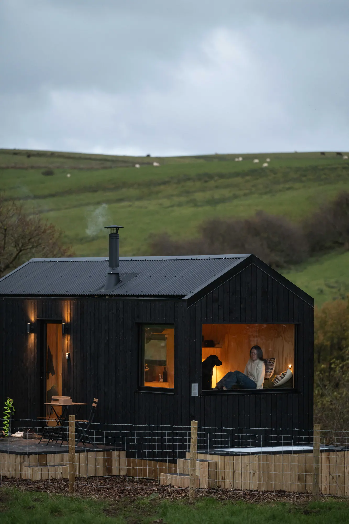 A black-timber Draco cabin in an open Cheshire field at dusk, warm light in the window