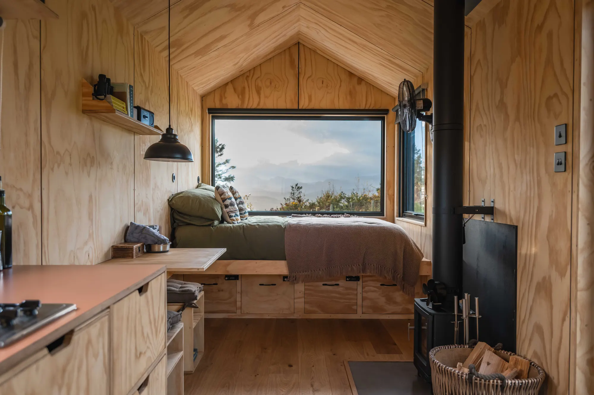 Inside the Draco cabin: a king bed by a picture window framing the hills, timber walls and a wood-burning stove