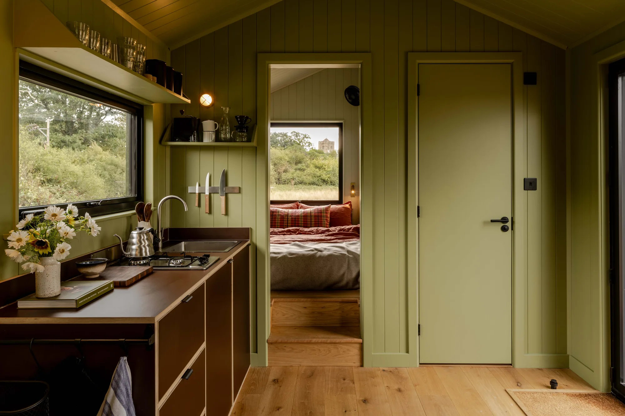 A sage-green cabin kitchen looking through to the bedroom, with a window onto the fields
