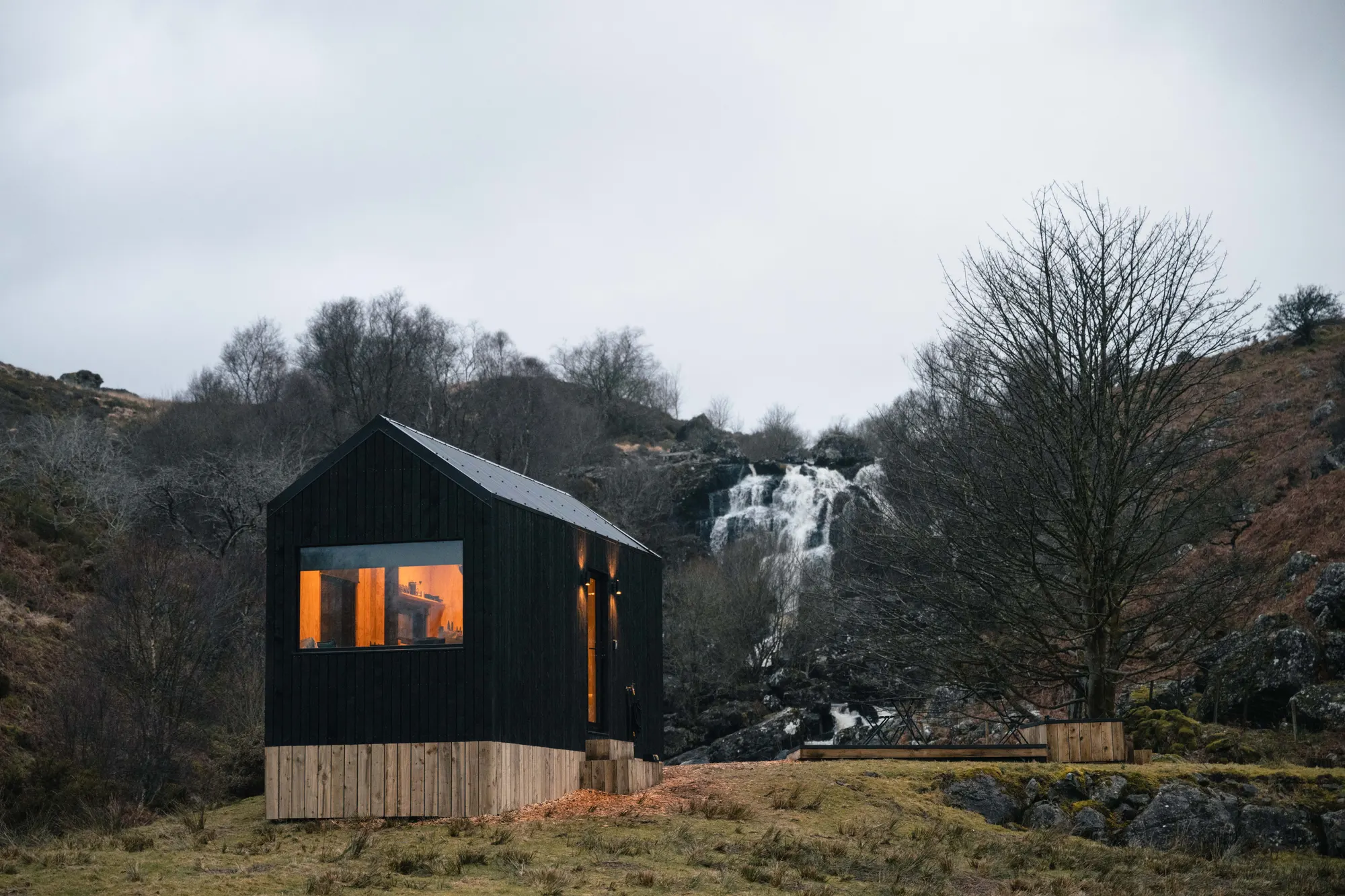 A black-timber Leo cabin beside a waterfall, warm light in the windows
