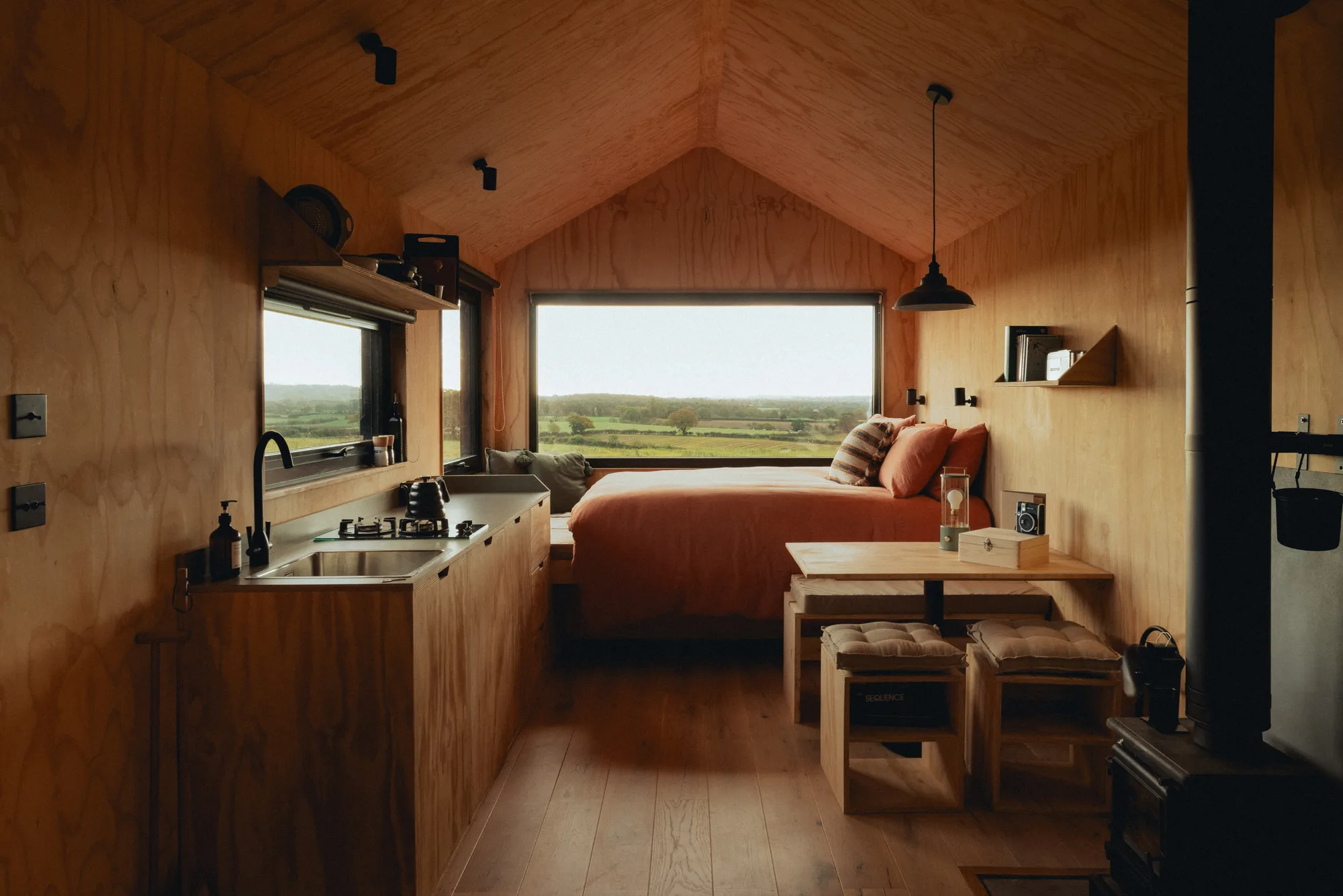 Inside the cabin: kitchen, dining table and bed beneath a wide window onto the fields, wood-burning stove alongside