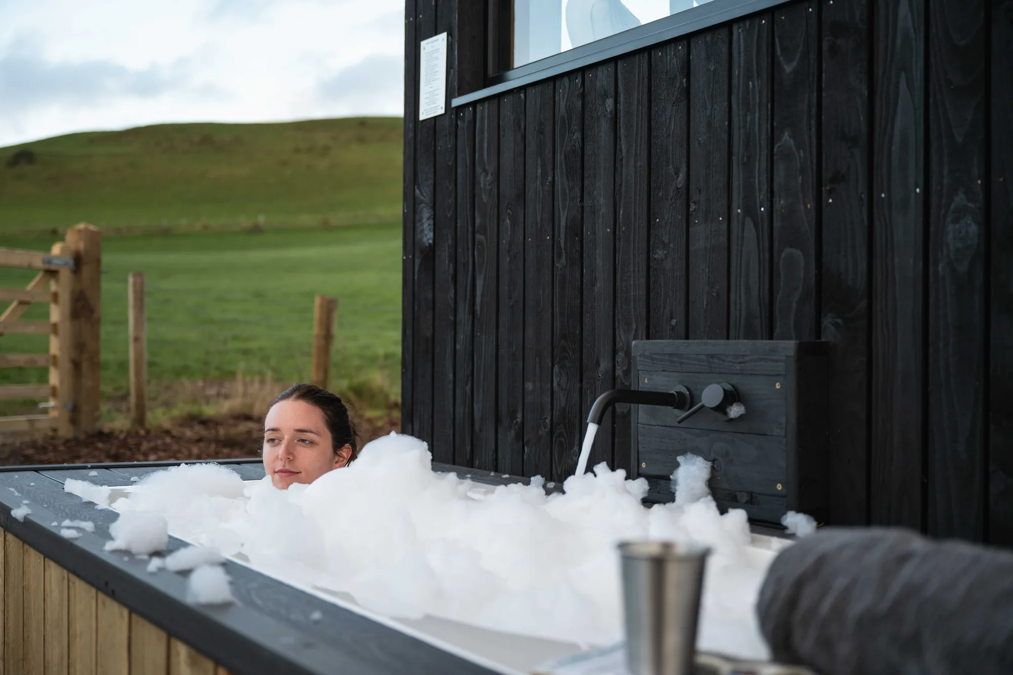 A guest soaking in the outdoor bath beside a black-timber Shacks cabin, green fields beyond