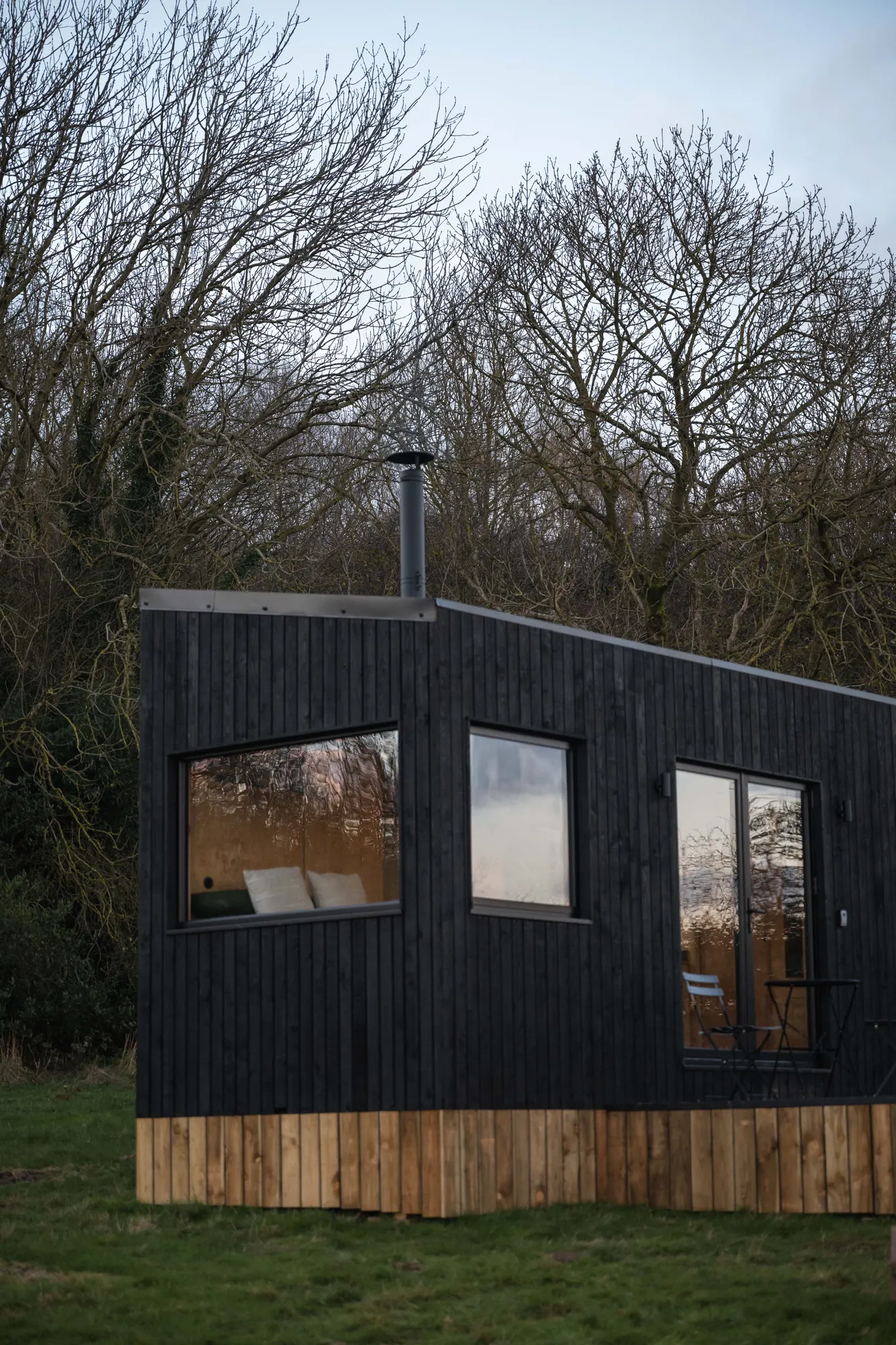 The Phoenix cabin in black timber with a larch base and lit windows, set against bare woodland