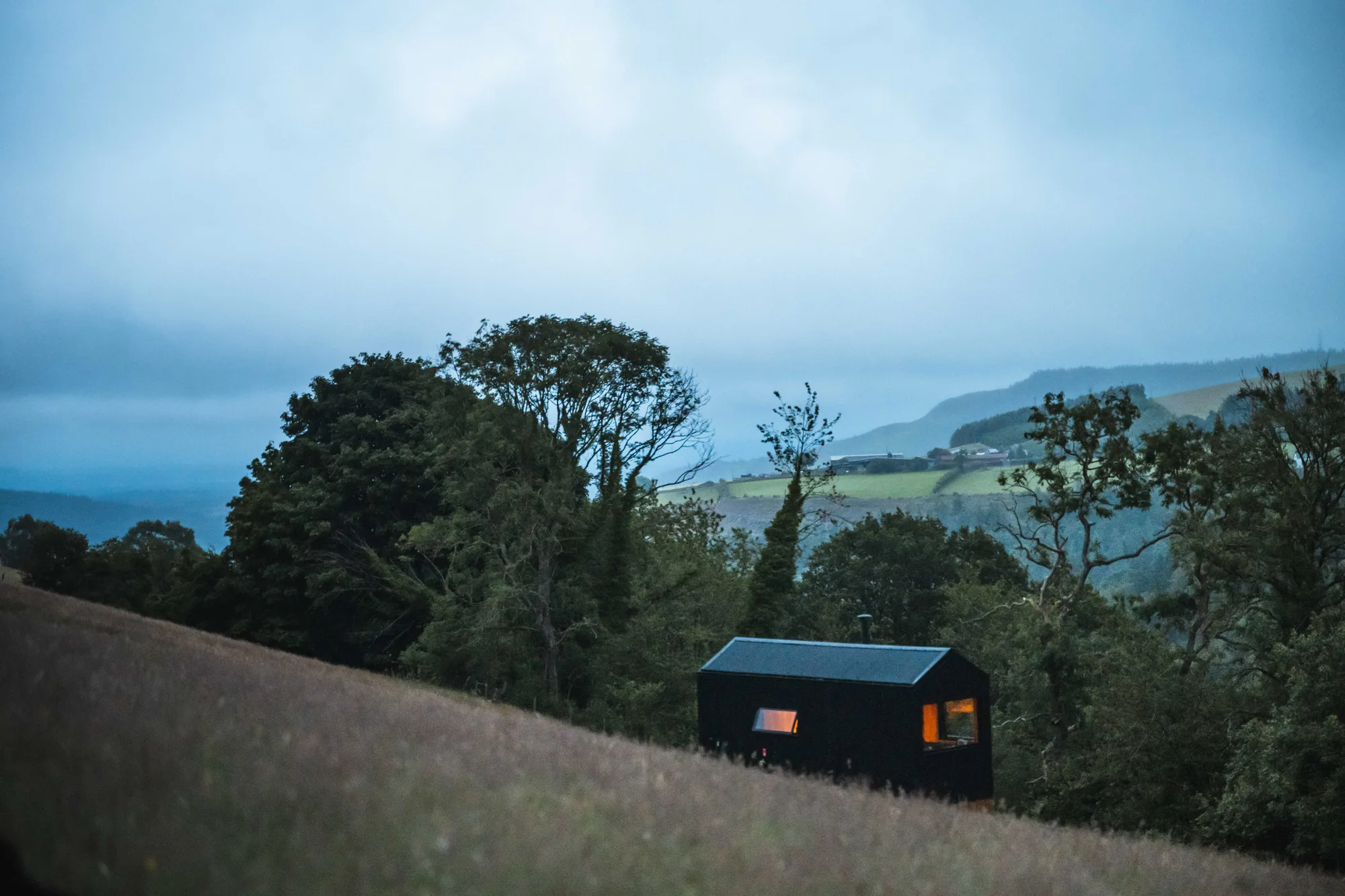 A black-timber Shacks cabin in a hillside meadow at dusk, looking out over the valley