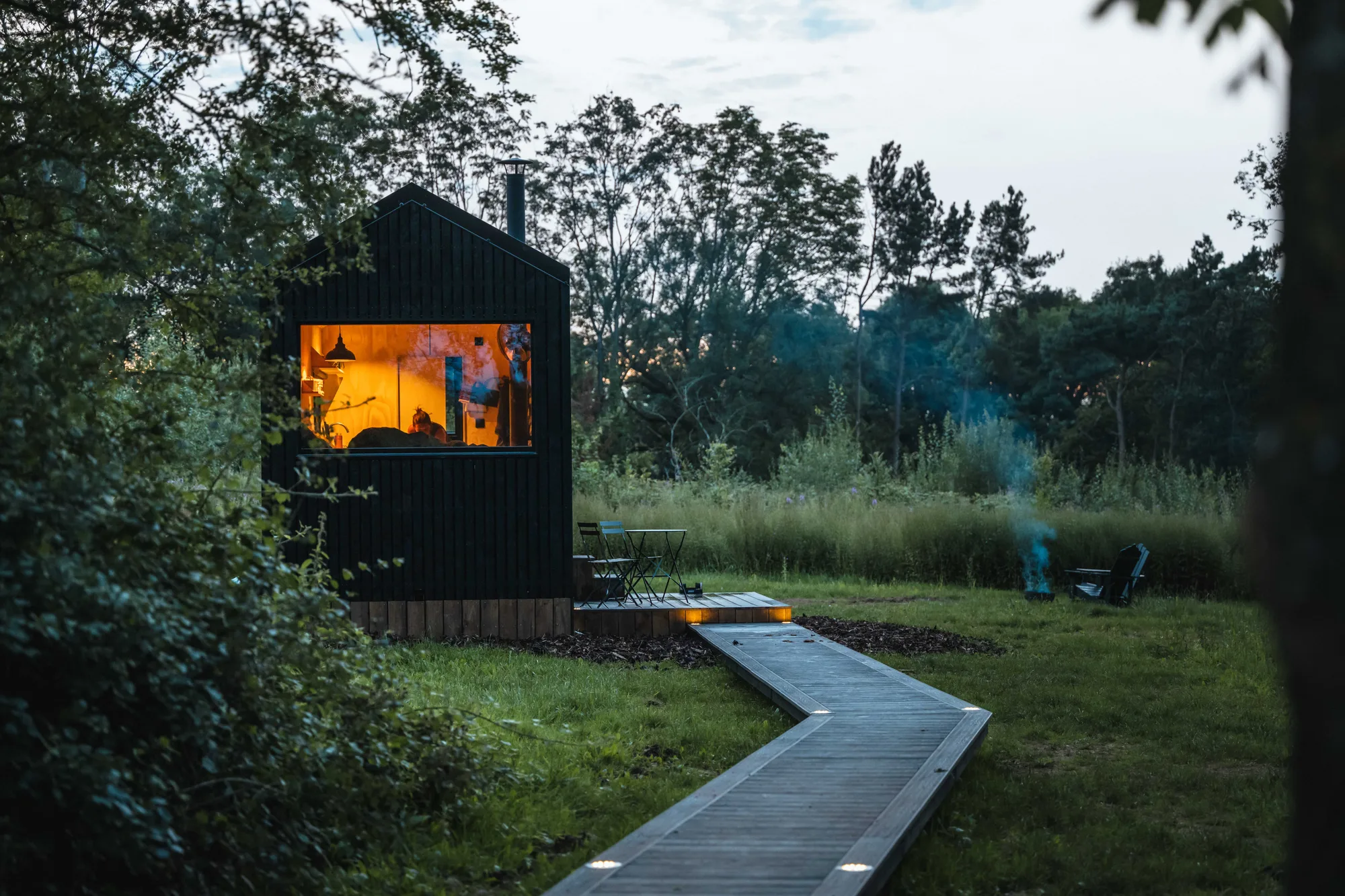 A black-timber Shacks cabin at dusk, warm light in the window and a lit boardwalk leading out to the fire pit
