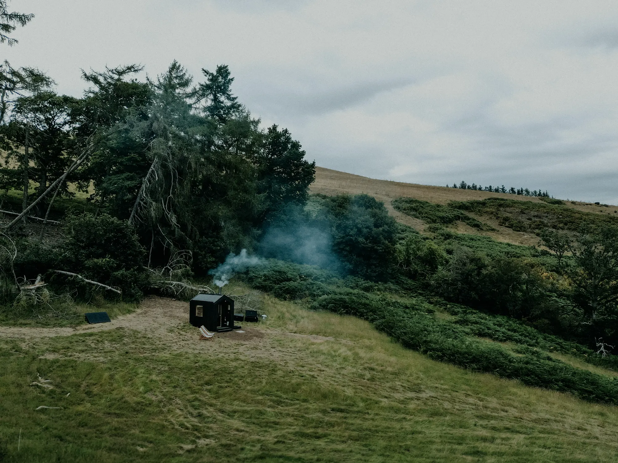 A black-timber Shacks sauna in a green field below wooded hills, woodsmoke rising
